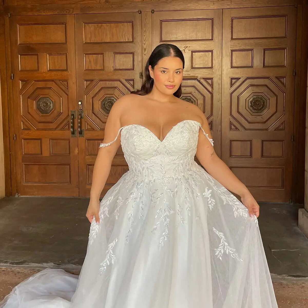 Latina bride wearing an off the shoulder wedding dress and standing in front of antique wooden doors. 