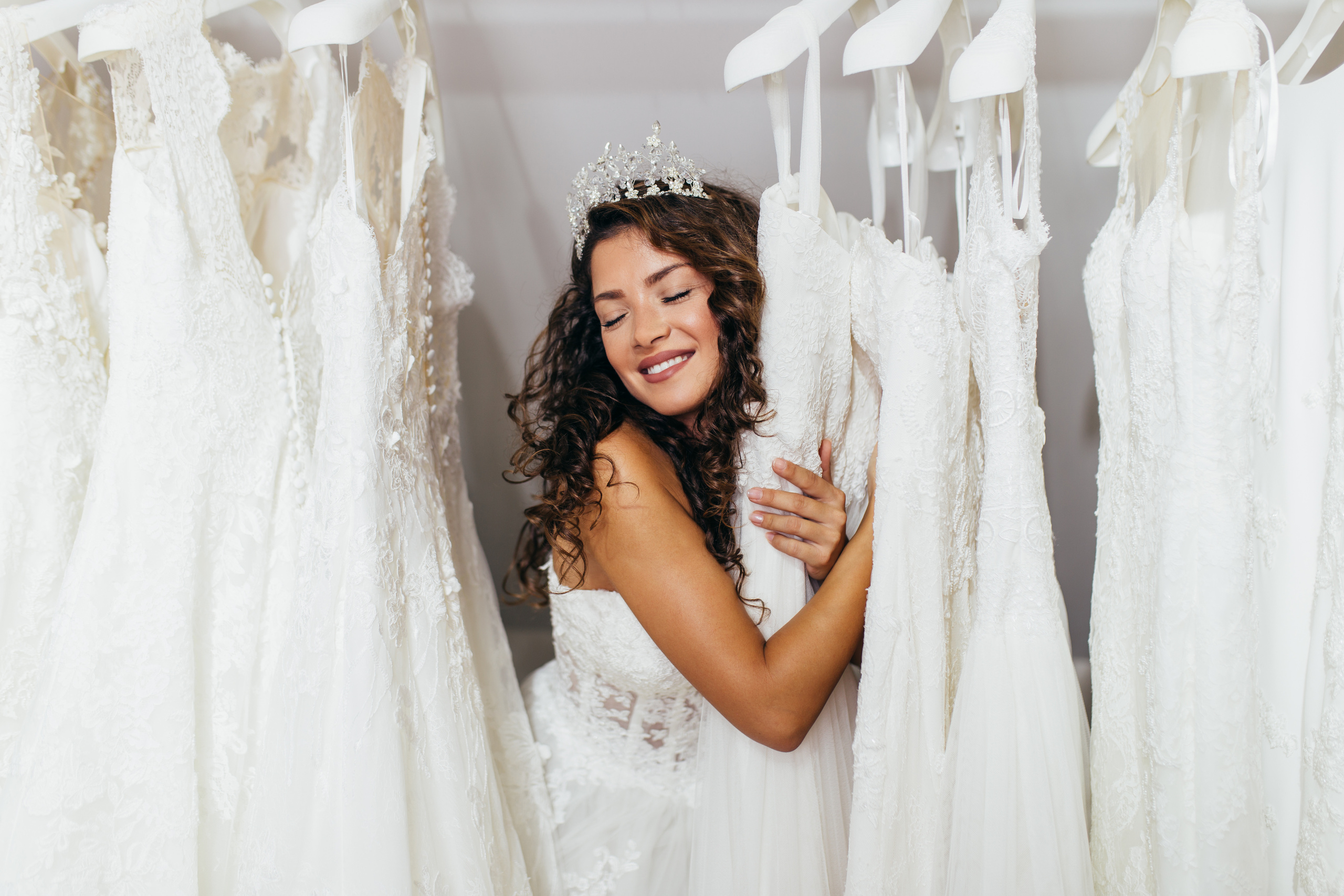 Young brunette wearing a wedding gown and crown, standing in a rack of wedding dresses. She is hugging the dress next to her and smiling with her eyes closed. 