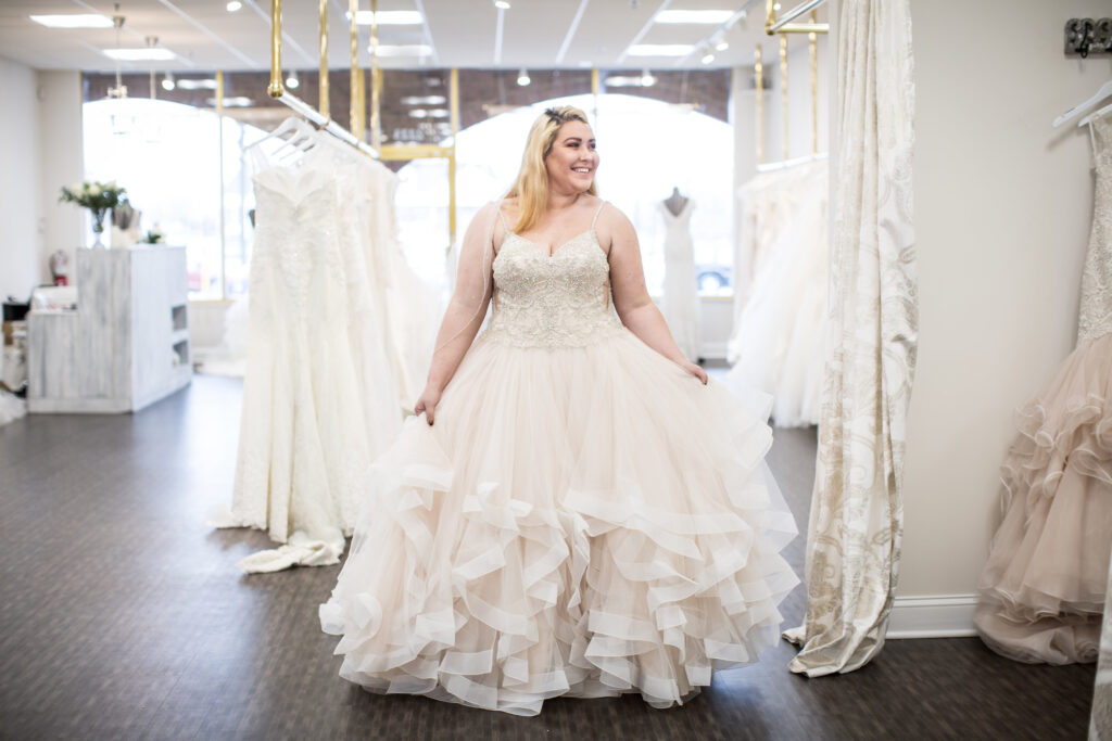 Bride to be standing in Vivid Bridal Boutique showing off a plus sized dress. The bodice has intricate beading and the skirt has lots of tulle layering.