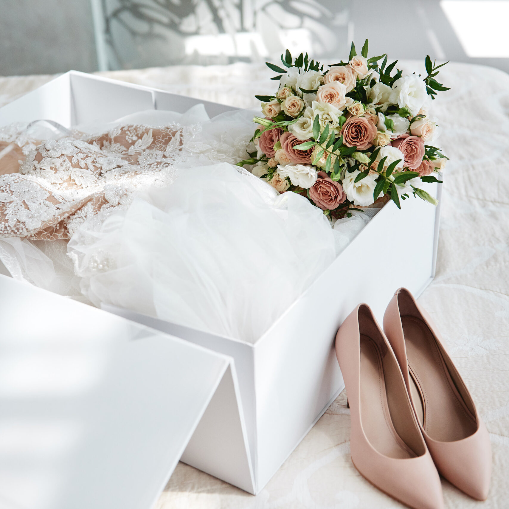 Luxury wedding dress in an open, white box with bouquet leaning on the side.  In front of the box is a pair of beige women's shoes.