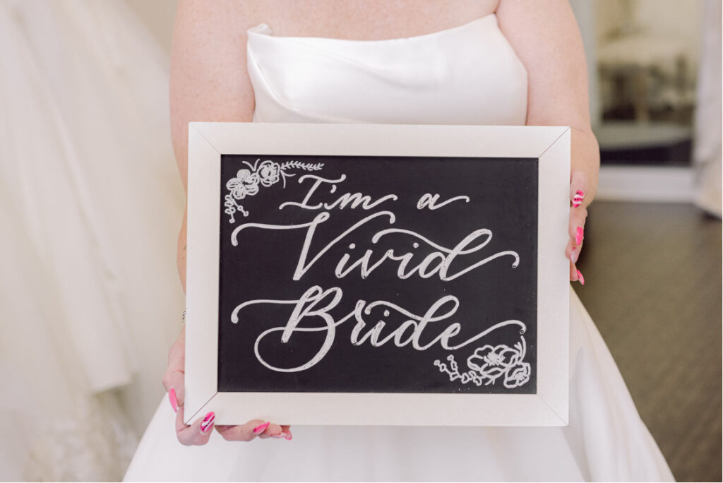 Bride with pink and white nails, wearing her wedding dress and holding a chalkboard sign saying "I'm a Vivid Bride" in front of her waist. 