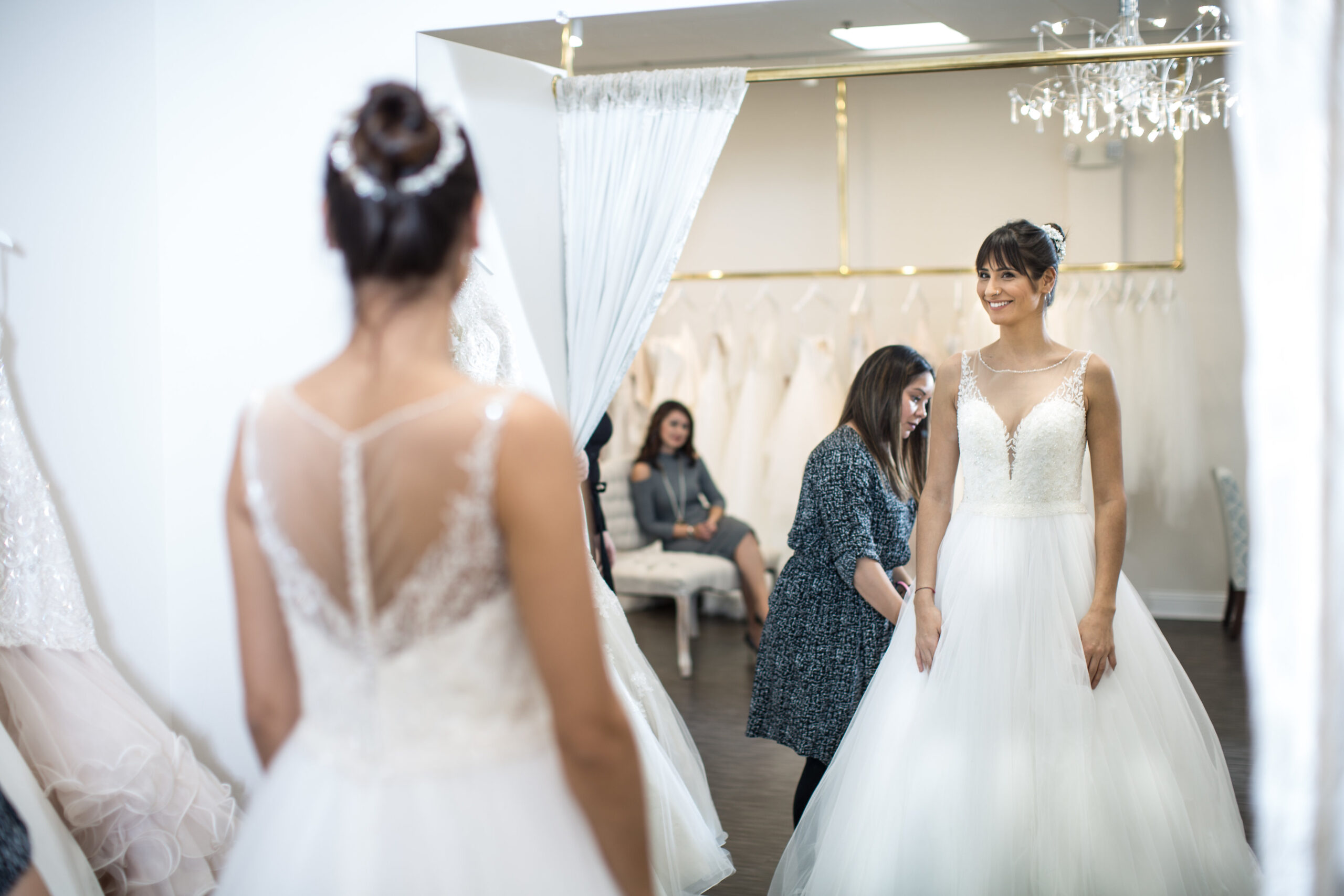 Bride to be standing in mirror looking at herself wearing a wedding gown while the sales person behind her adjusts the train.