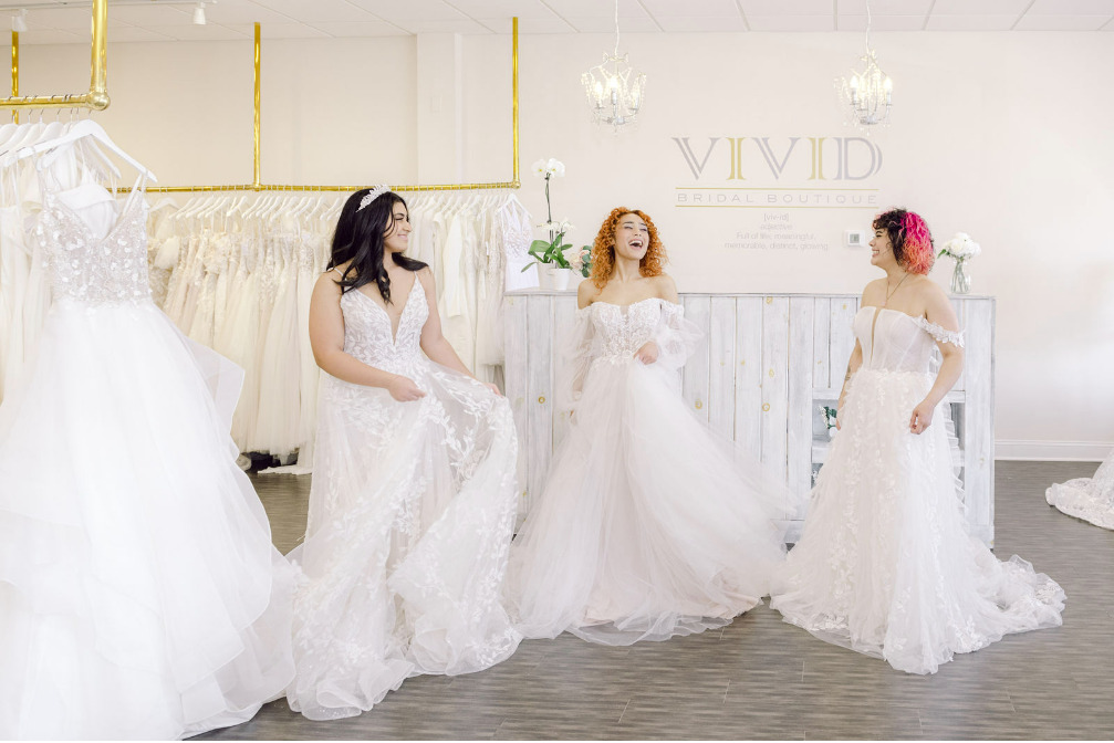 Three brides-to-be standing in the lobby of Vivid Bridal Boutique. All three are wearing wedding gowns and laughing together.