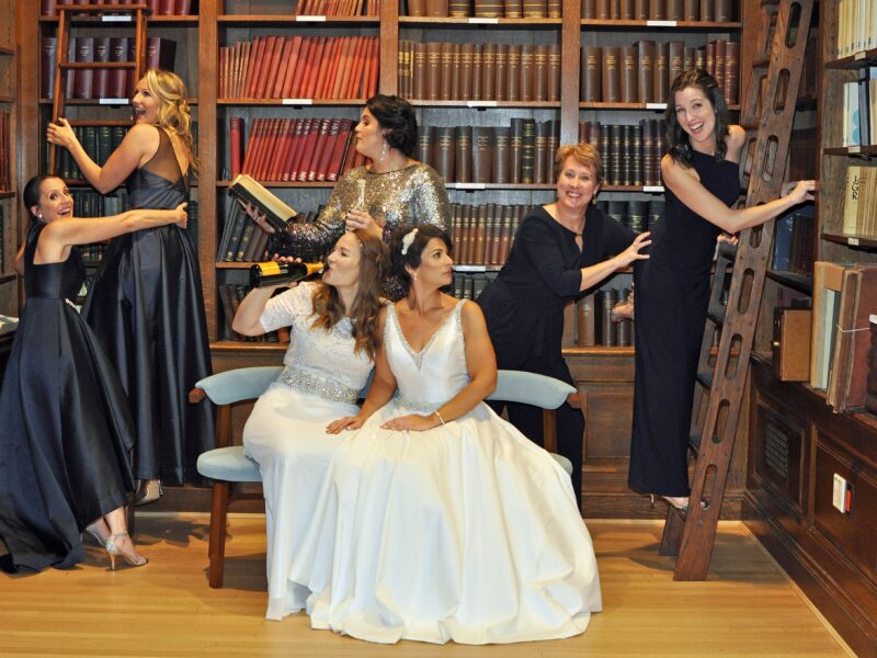 Teacher Bride-to-Be sitting on a bench in a library with her bridesmaids surrounding her.